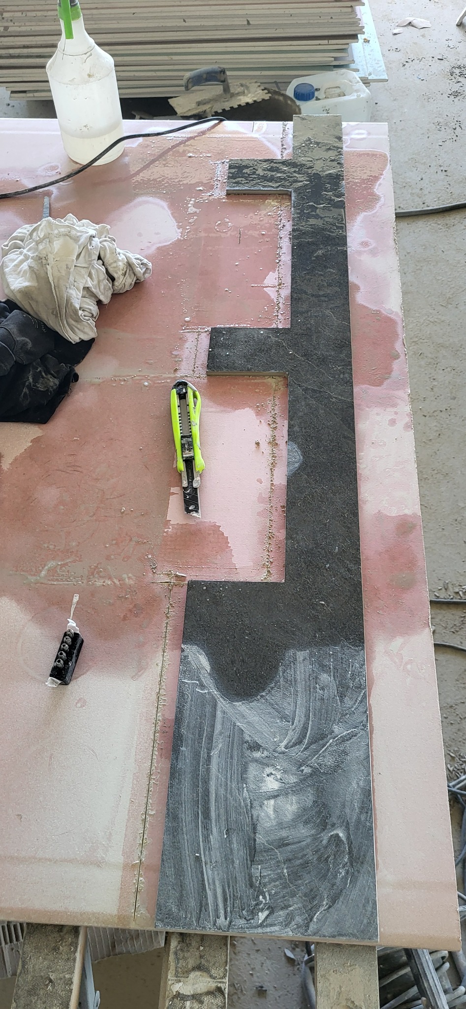 Przygotowanie płytek: docinanie ciemnej płytki na różowej płycie, widoczny nóż do cięcia, klej, rękawice i butelka z płynem. Blat roboczy w trakcie prac glazurniczych.