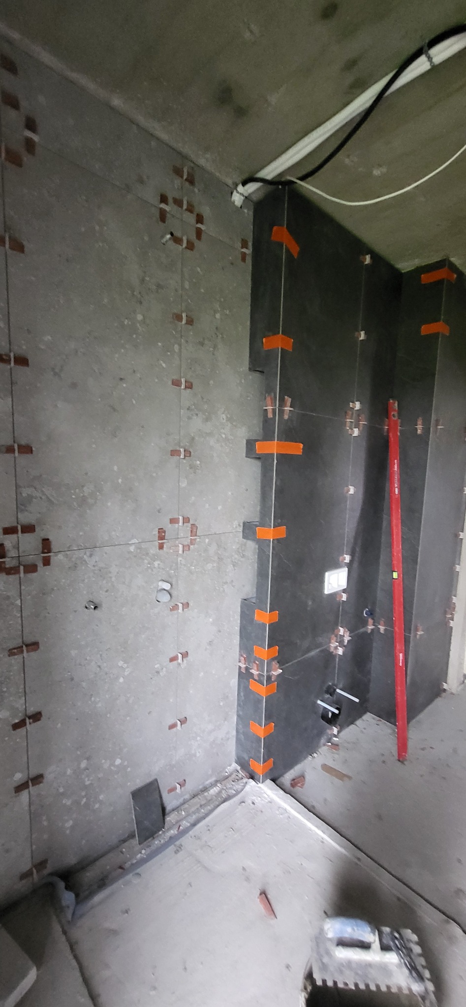 Częściowo wykończona ściana z ciemnych płytek w łazience, z pomarańczowymi krzyżykami dystansowymi i poziomnicą. Widoczne instalacje i surowy beton.