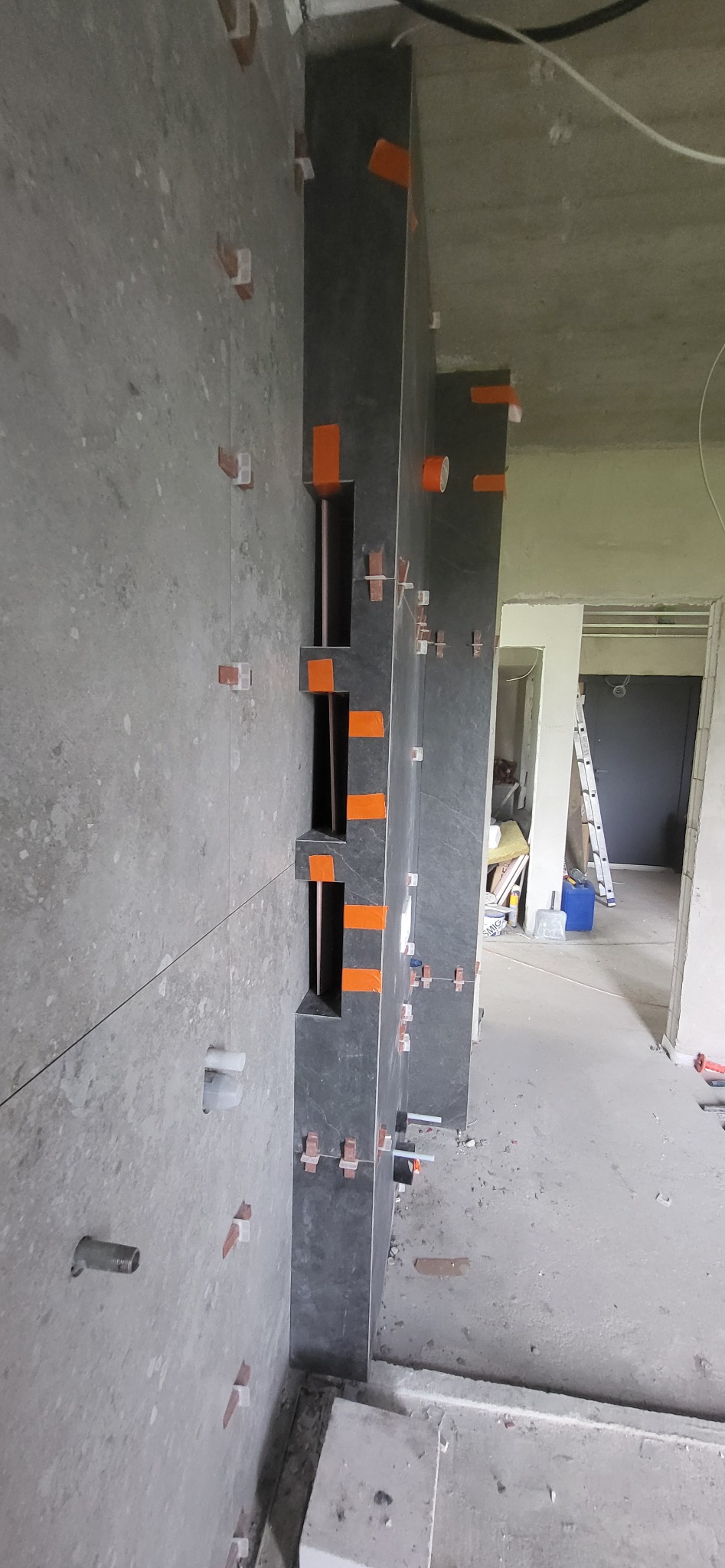 Montaż ciemnych płytek z wnękami na ścianie, widoczne krzyżyki dystansowe i taśma. W tle surowy stan pomieszczenia z drabiną.