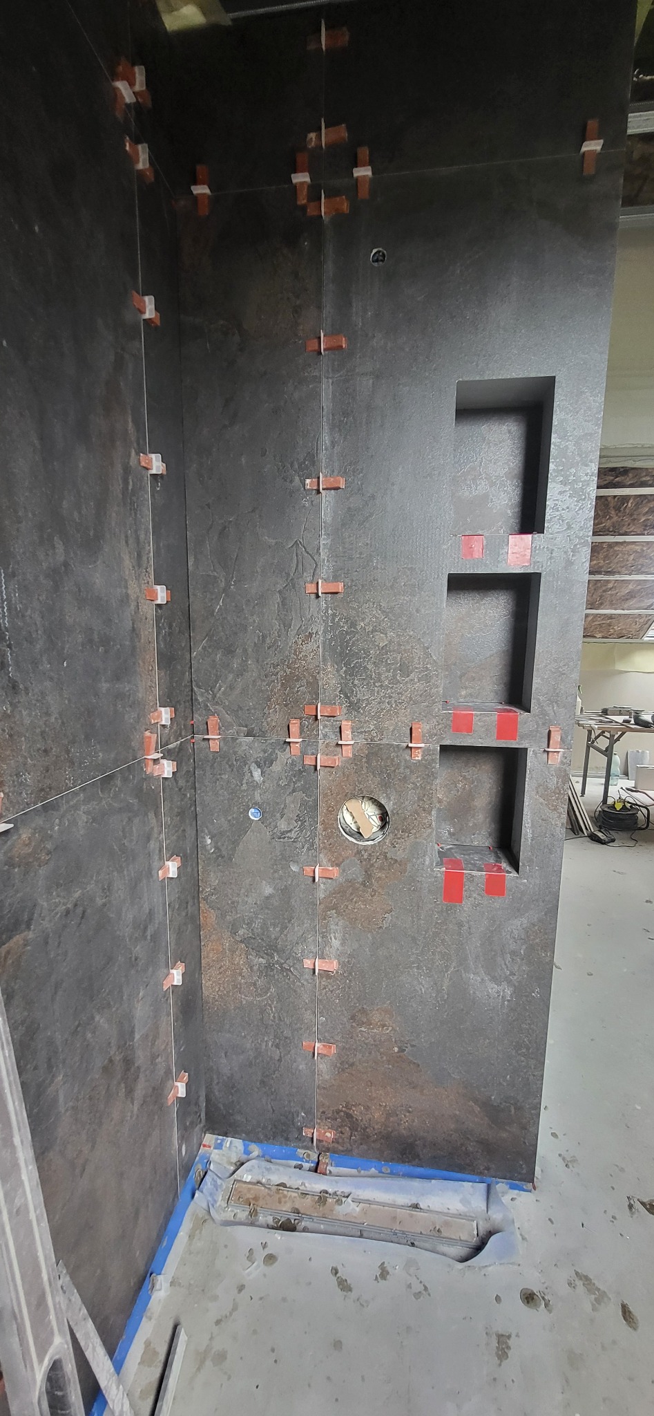 Ciemne płytki imitujące kamień, ułożone w rogu łazienki z wnękami. Widoczne krzyżyki dystansowe i pionowe sznurki, sugerujące etap prac glazurniczych.