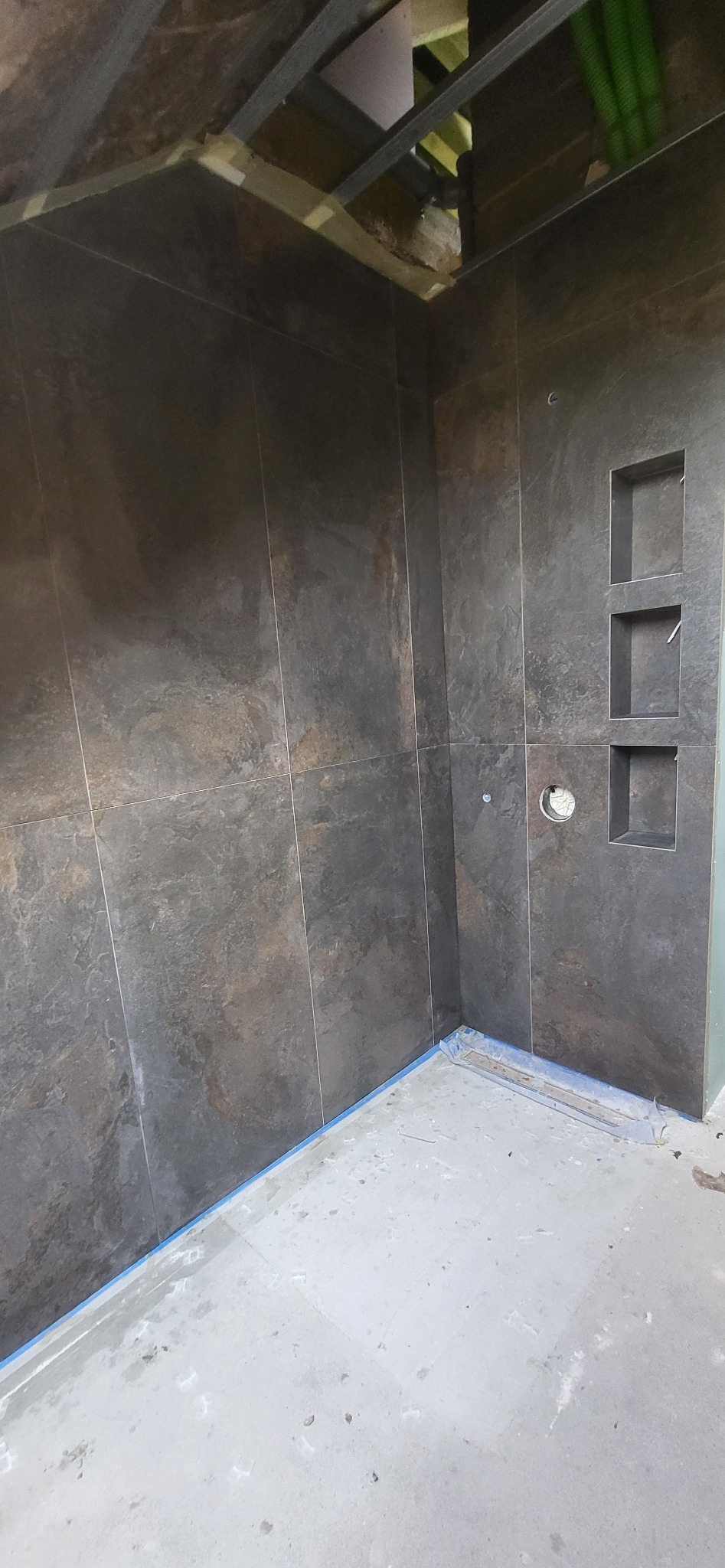 Wykończona ściana w łazience z ciemnymi płytkami imitującymi kamień, wnękami i otworem na armaturę. Widoczny fragment sufitu z instalacją.