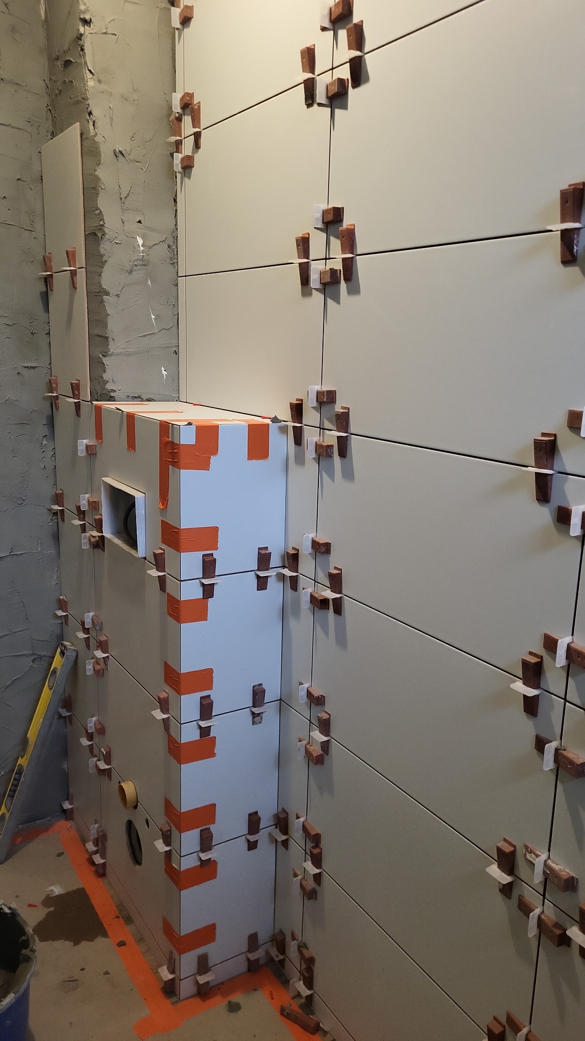 Ściana z jasnych, kwadratowych płytek, w trakcie montażu, z użyciem krzyżyków dystansowych. Widoczny fragment ściany z surowym tynkiem i obudowa z płyt.