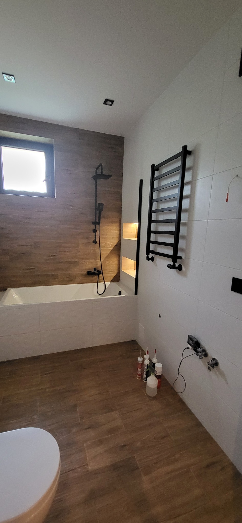 Nowoczesna łazienka z wanną, brązowymi płytkami imitującymi drewno i czarnymi akcentami: grzejnik, armatura. Widoczne materiały montażowe. Minimalistyczny design.