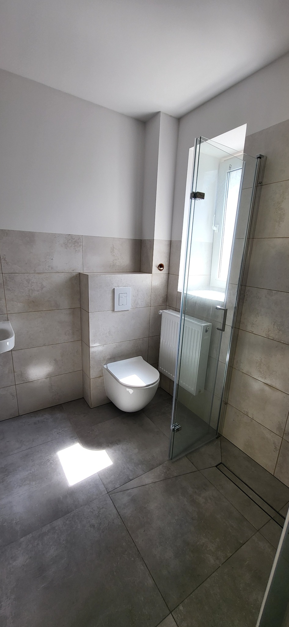 Nowoczesna łazienka po remoncie: beżowe płytki ścienne, szare płytki podłogowe, podwieszana toaleta, szklana kabina prysznicowa i grzejnik. Minimalistyczny design.