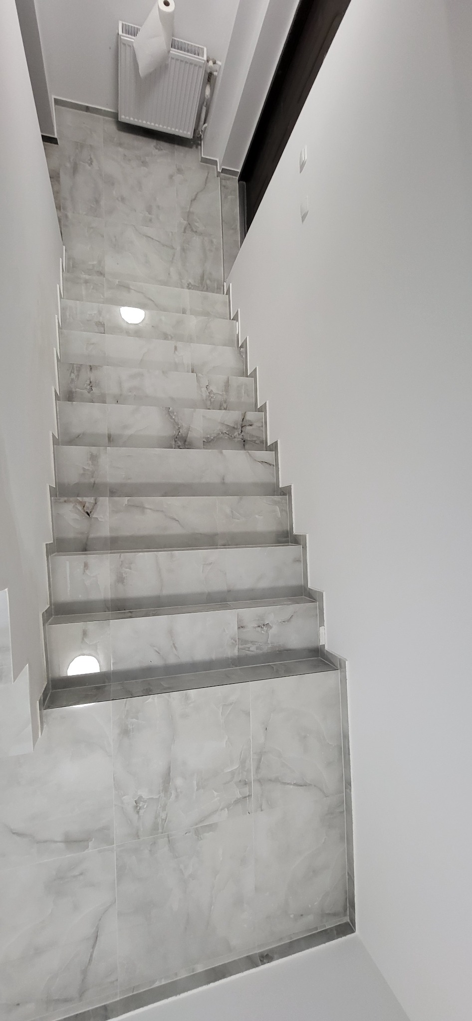 Wykończone schody z szarych płytek imitujących marmur, z oświetleniem punktowym w stopniach. Widok z góry, białe ściany i grzejnik w tle.