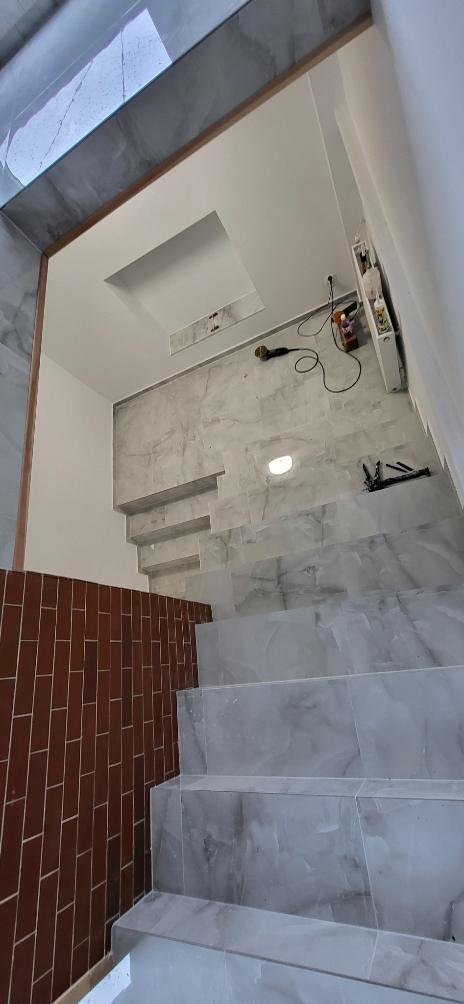 Wykończone schody z szarych płytek imitujących marmur, brązowa ściana z płytek cegiełek, sufit z podwieszanym elementem. Widoczne narzędzia glazurnicze.