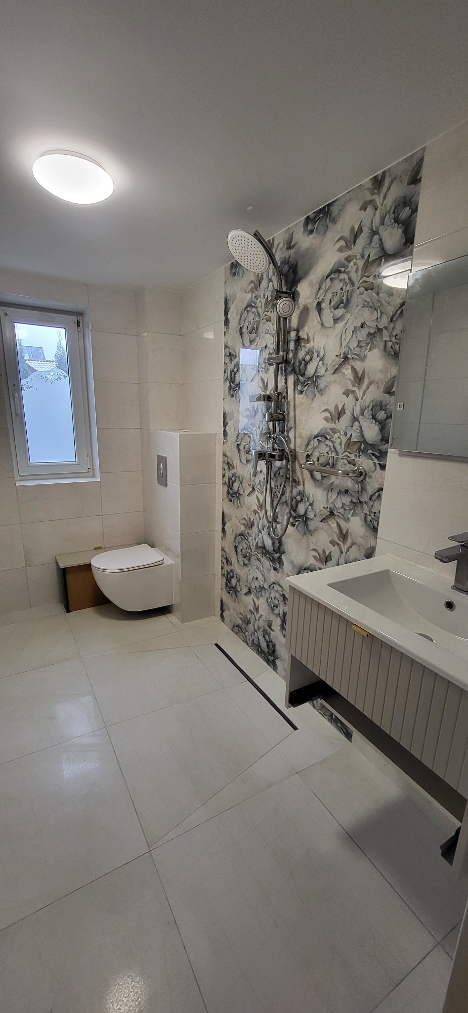 Wykończona łazienka z białymi płytkami, wiszącą toaletą i kabiną prysznicową z deszczownicą. Ściana akcentowa z motywem kwiatowym. Nowoczesna armatura i oświetlenie.