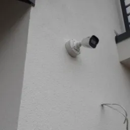 Biała kamera monitoringu zamontowana na elewacji budynku. Widoczne przewody, orynnowanie i fragment balkonu. Kamera skierowana w lewo.