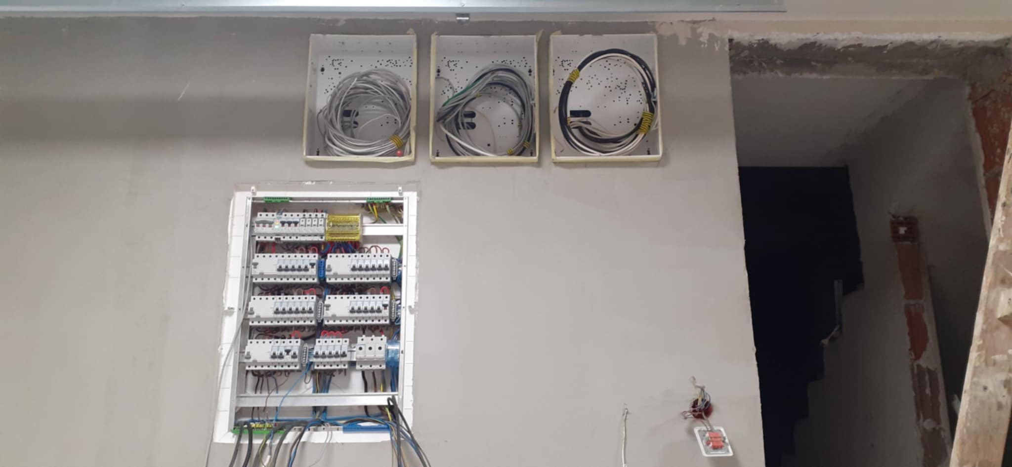 Rozdzielnica elektryczna z bezpiecznikami i okablowaniem w trzech puszkach na ścianie podczas prac wykończeniowych, widoczne przewody i elementy instalacji.
