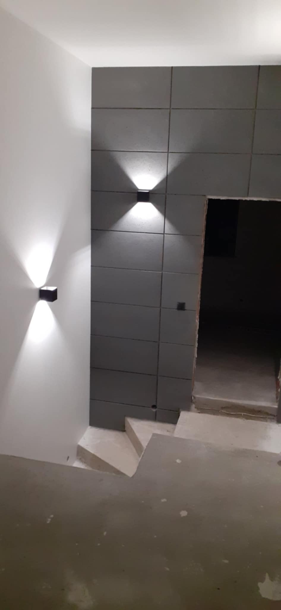 Nowoczesne oświetlenie schodów: dwie czarne lampy ścienne emitują światło w górę i w dół, oświetlając białą ścianę i szare płytki. Minimalistyczny design.