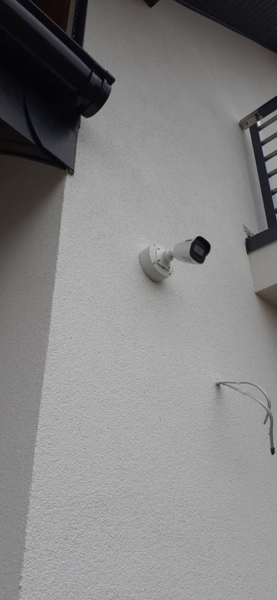 Biała kamera monitoringu zamontowana na elewacji budynku. Widoczne przewody, orynnowanie i fragment balkonu. Kamera skierowana w lewo.