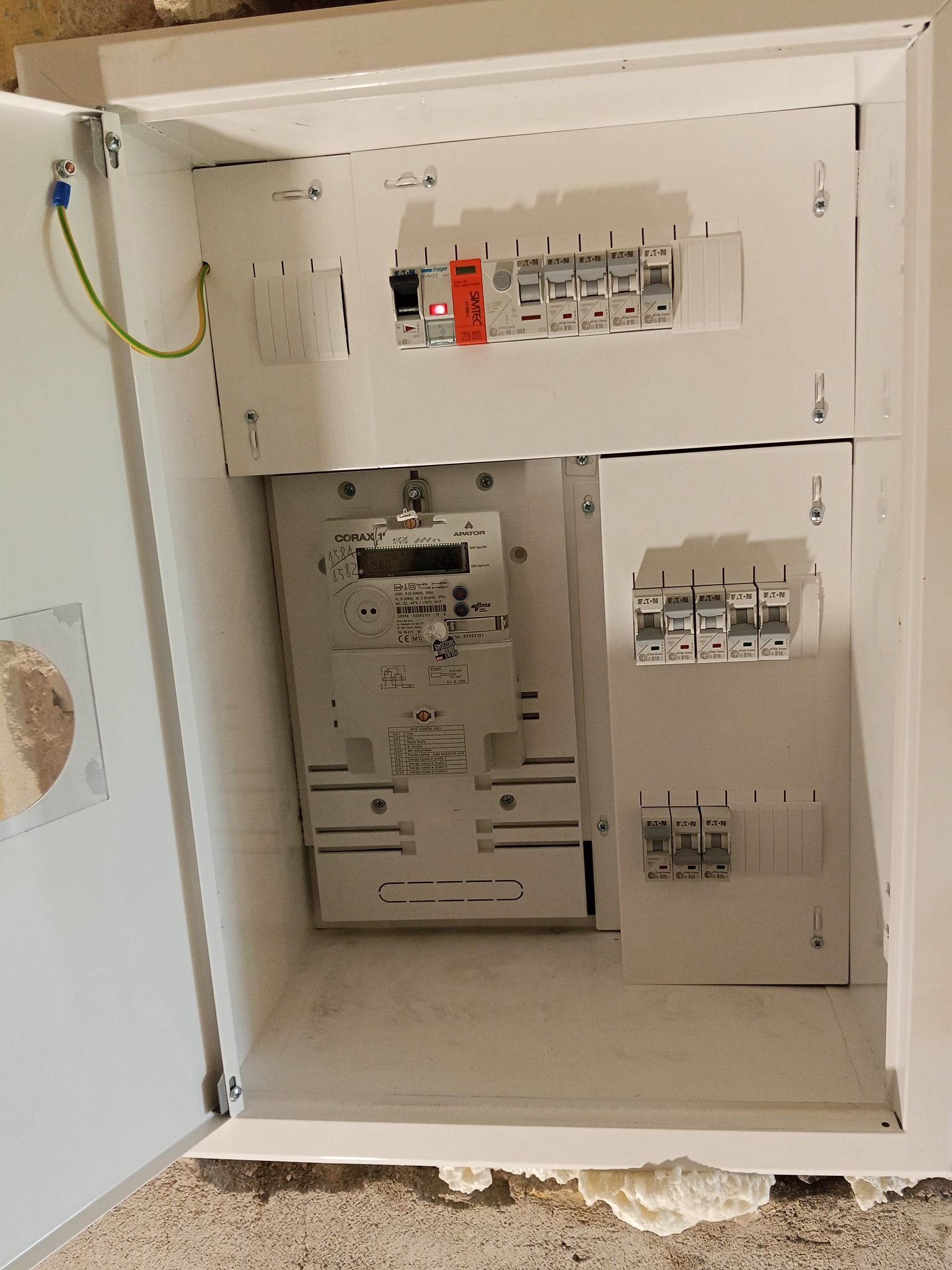 Otwarta, biała skrzynka elektryczna z zamontowanym licznikiem i bezpiecznikami Eaton. Widoczne kable i elementy instalacji. Detal złącza uziemiającego na drzwiach.