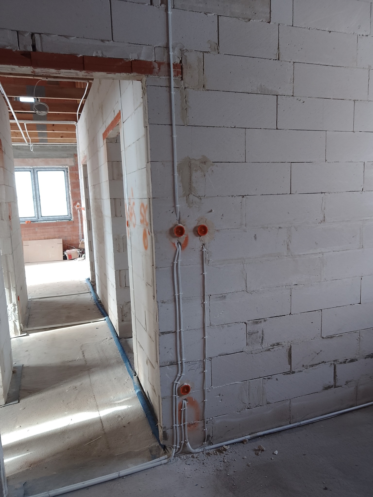 Surowy mur z instalacją elektryczną: kable w peszlach prowadzone po ścianie, widoczne pomarańczowe puszki instalacyjne. W tle surowy stan budynku, okno.