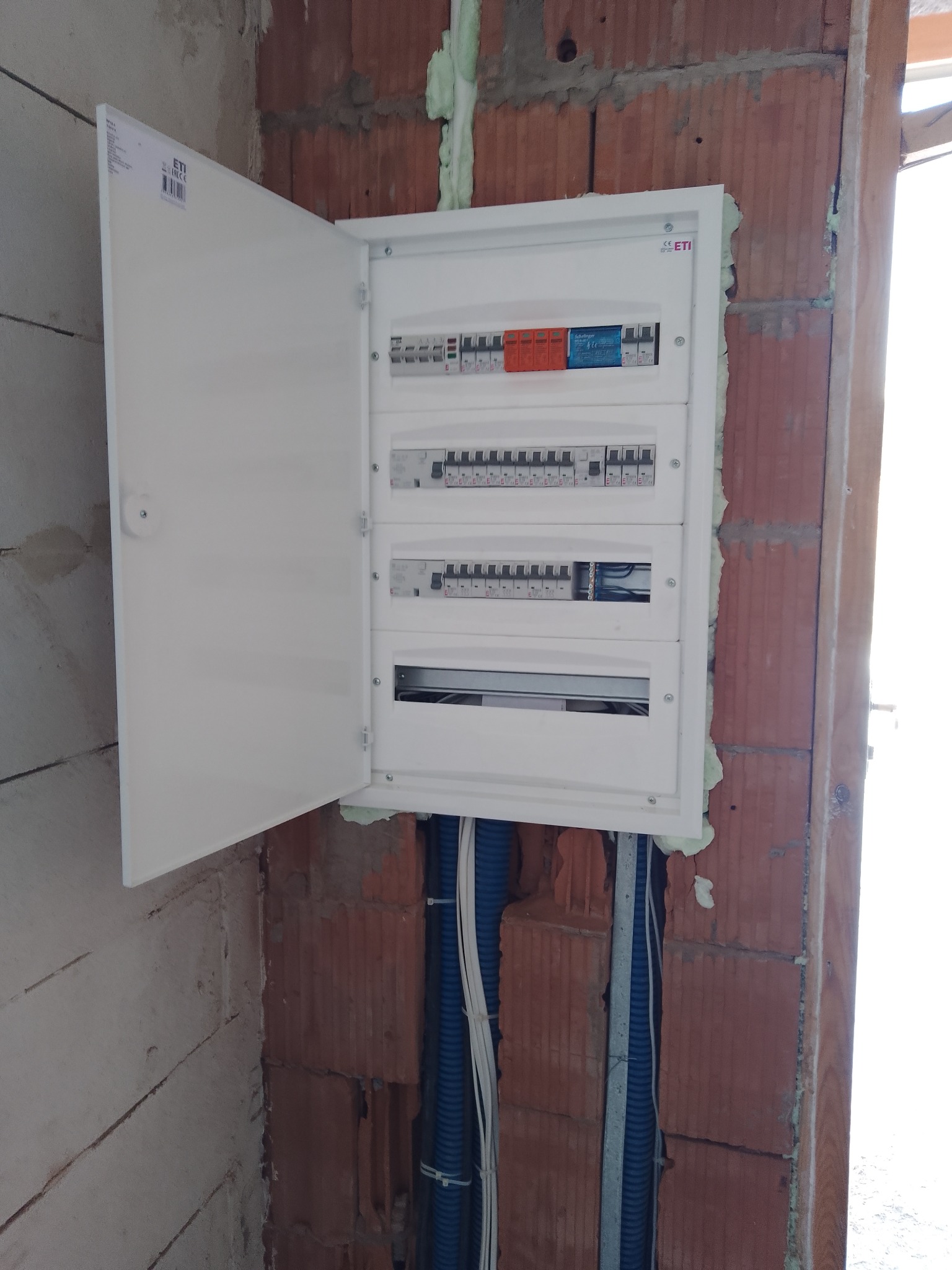 Otwarta skrzynka elektryczna z zainstalowanymi bezpiecznikami, zamontowana na ceglanej ścianie, widoczne niebieskie rury instalacyjne.