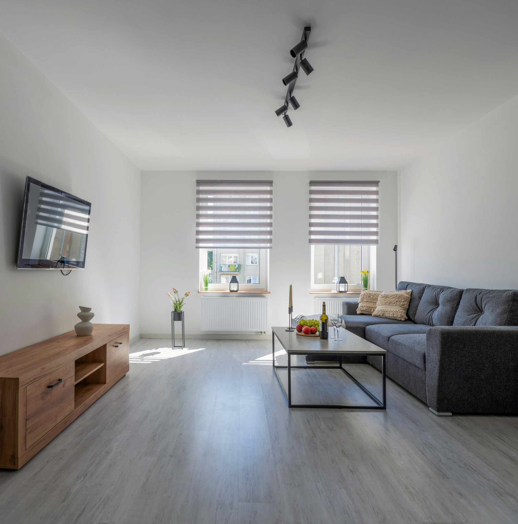 Jasne, przestronne wnętrze salonu z szarym narożnikiem, drewnianą szafką RTV i podłogą imitującą drewno, oświetlone światłem dziennym wpadającym przez okna z roletami dzień-noc.