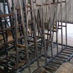 Konstrukcja z metalowych profili spawanych, tworząca ramy z uchwytami, prawdopodobnie do transportu elementów szklanych lub kamiennych, widoczne spawy, surowe wykończenie, w tle brama garażowa.