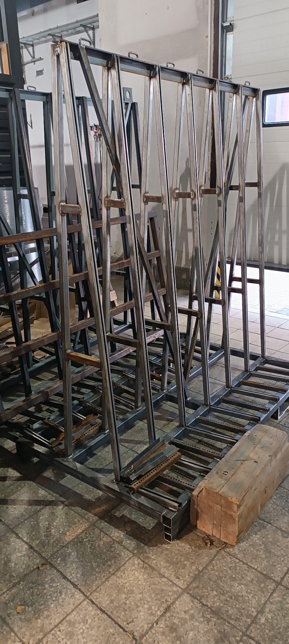 Konstrukcja z metalowych profili spawanych, tworząca ramy z uchwytami, prawdopodobnie do transportu elementów szklanych lub kamiennych, widoczne spawy, surowe wykończenie, w tle brama garażowa.