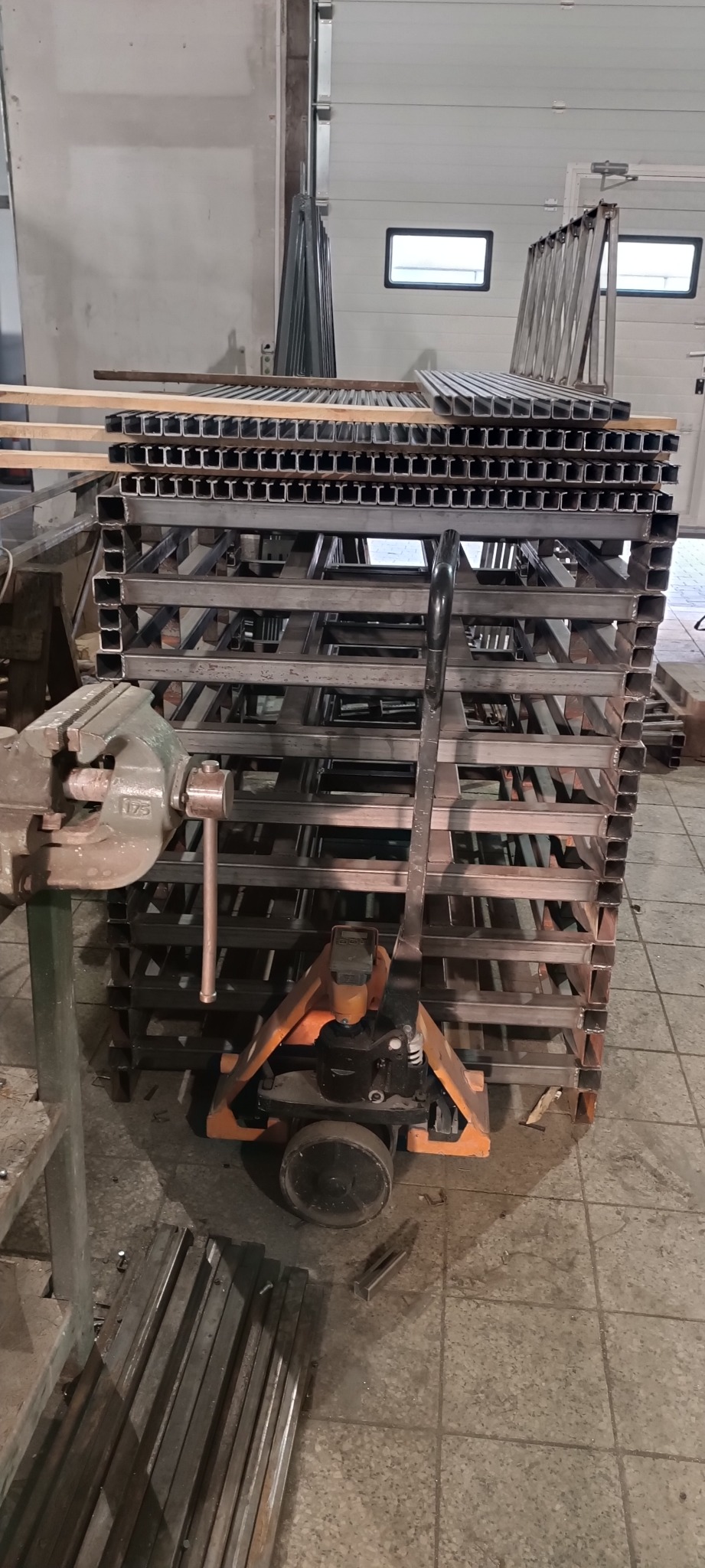 Stos kwadratowych profili metalowych ułożonych na paleciaku, widoczna prasa śrubowa w warsztacie.