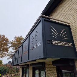 Krycmet Ruslan Kuraka - Czarna, metalowa balustrada balkonowa z ażurowym wzorem roślinnym, zamontowana na ceglanym budynku. Widok z dołu, jesienne drzewa w tle.