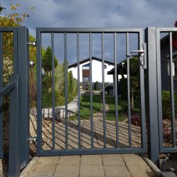 Krycmet Ruslan Kuraka - Nowoczesna, grafitowa brama wjazdowa z pionowymi szczebelkami, widziana z bliska, z domem i zielenią w tle. Minimalistyczny design, równomierne oświetlenie.