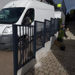 Krycmet Ruslan Kuraka - Nowoczesne, grafitowe ogrodzenie posesji z pionowych sztachet, zamontowane na betonowym murku. W tle biały bus i schody z pracownikiem w trakcie sadzenia roślin.