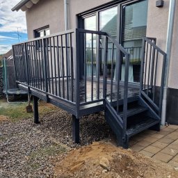 Krycmet Ruslan Kuraka - Nowoczesny, czarny taras z metalową balustradą i schodami, przylegający do domu. Widoczne kamienie i kostka brukowa wokół tarasu. Ujęcie z dołu, podkreślające konstrukcję.