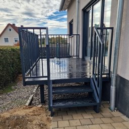 Krycmet Ruslan Kuraka - Metalowa konstrukcja tarasu z balustradą i schodami, pomalowana na ciemny kolor, przylegająca do budynku. Widoczne elementy otoczenia: trawnik, kostka brukowa i fragment nieba.