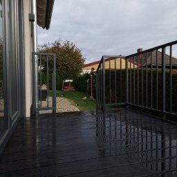 Krycmet Ruslan Kuraka - Ciemna, metalowa balustrada balkonowa z furtką, widok z góry na mokrą podłogę balkonu, w tle osoba z łopatą w ogrodzie.