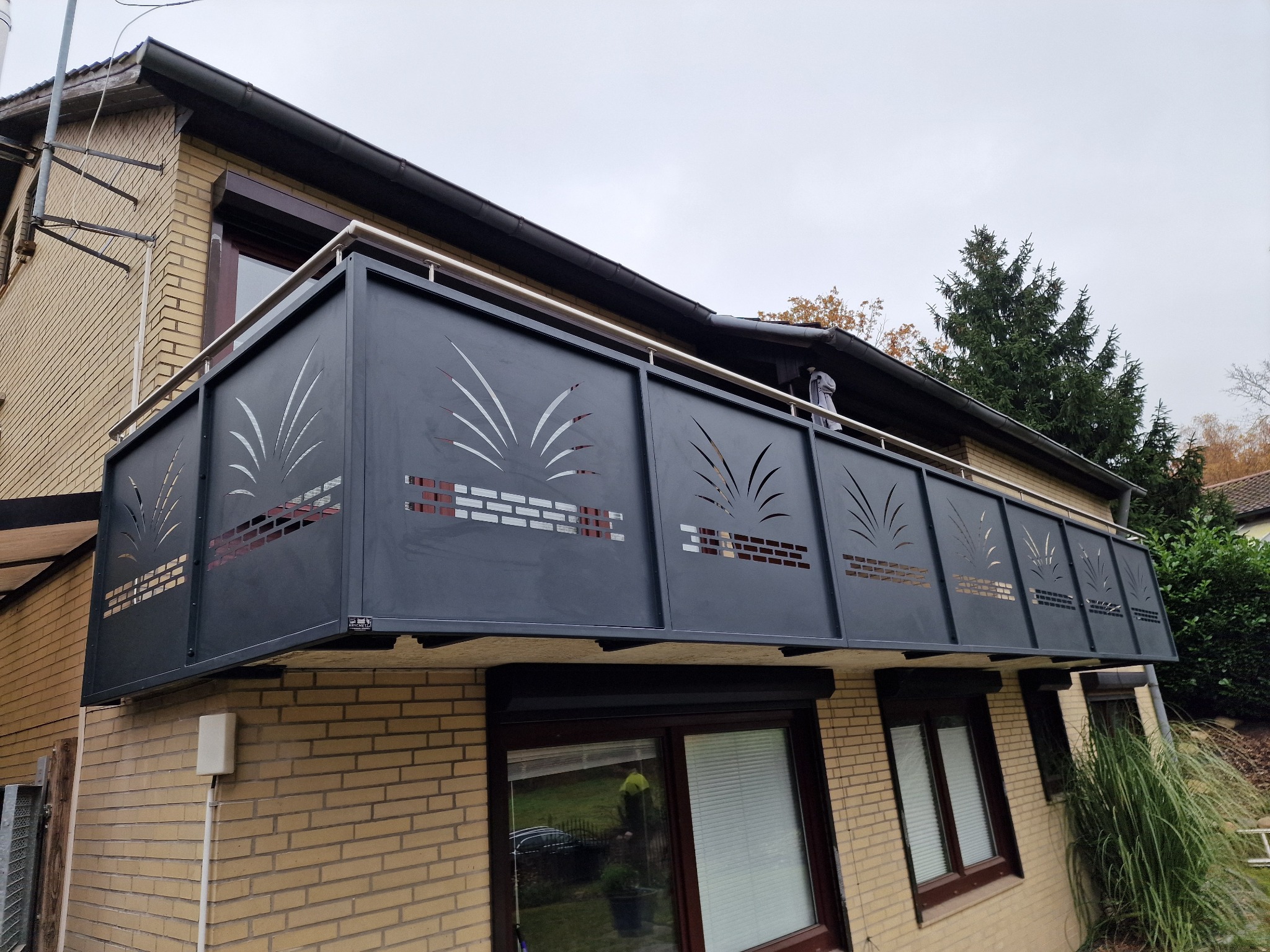 Nowoczesna, metalowa balustrada balkonowa z laserowo wycinanymi wzorami trawy, zamontowana na ceglanym budynku. Szara kolorystyka, stalowa poręcz, widok z dołu.