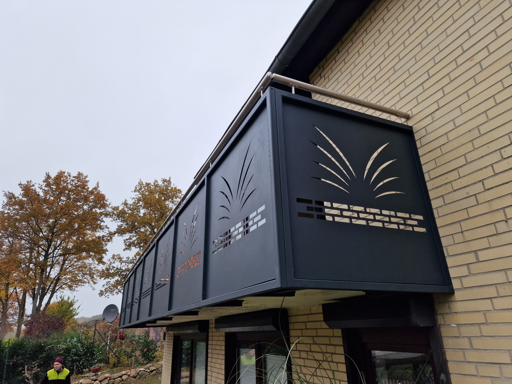 Czarna, metalowa balustrada balkonowa z ażurowym wzorem roślinnym, zamontowana na ceglanym budynku. Widok z dołu, jesienne drzewa w tle.