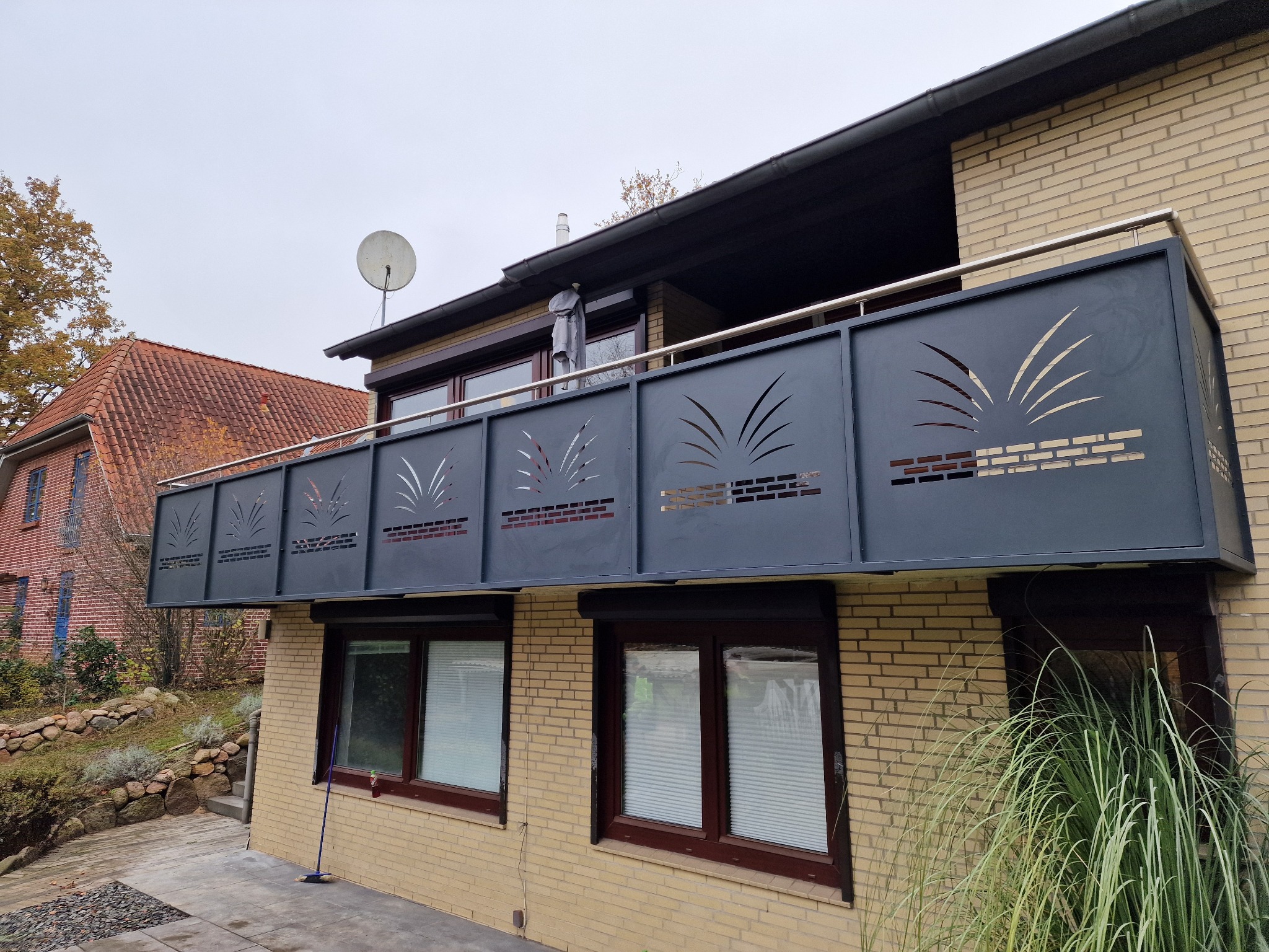 Nowoczesna balustrada balkonowa z metalu, z wycinanym wzorem roślinnym, zamontowana na balkonie budynku z żółtej cegły. Widok z dołu, w tle budynek z czerwoną dachówką.