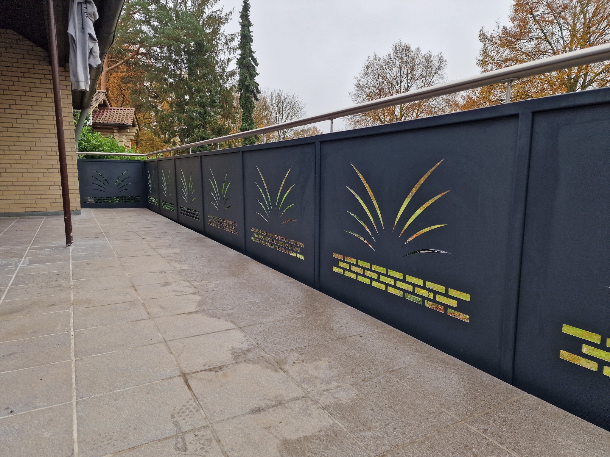 Nowoczesna, ażurowa balustrada balkonowa w Dębnie. Czarne panele z motywem roślinnym i cegieł, stalowa poręcz. Widok z boku, płytki na balkonie.