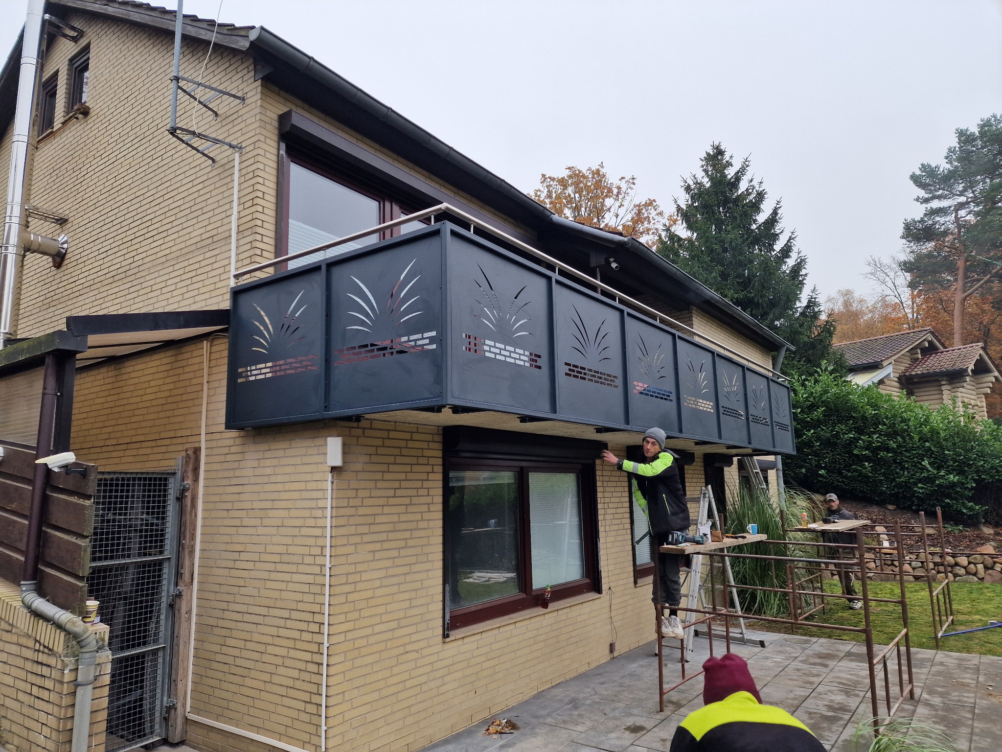 Montaż czarnej balustrady balkonowej z ażurowym wzorem na dwupiętrowym domu z cegły. Pracownik na rusztowaniu przytwierdza element. Widoczne narzędzia i fragment ogrodu.