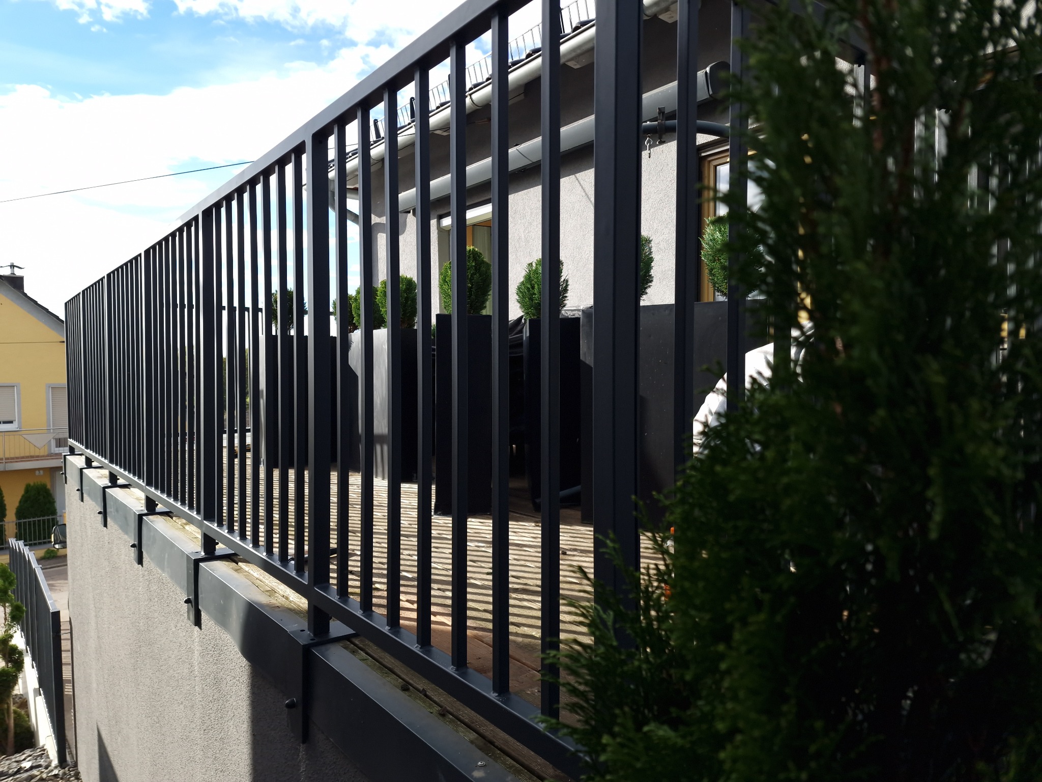 Nowoczesna, czarna balustrada balkonowa z pionowymi szczeblami, zamontowana na szarym murku. W tle widoczne tuje w donicach i fragment budynku.