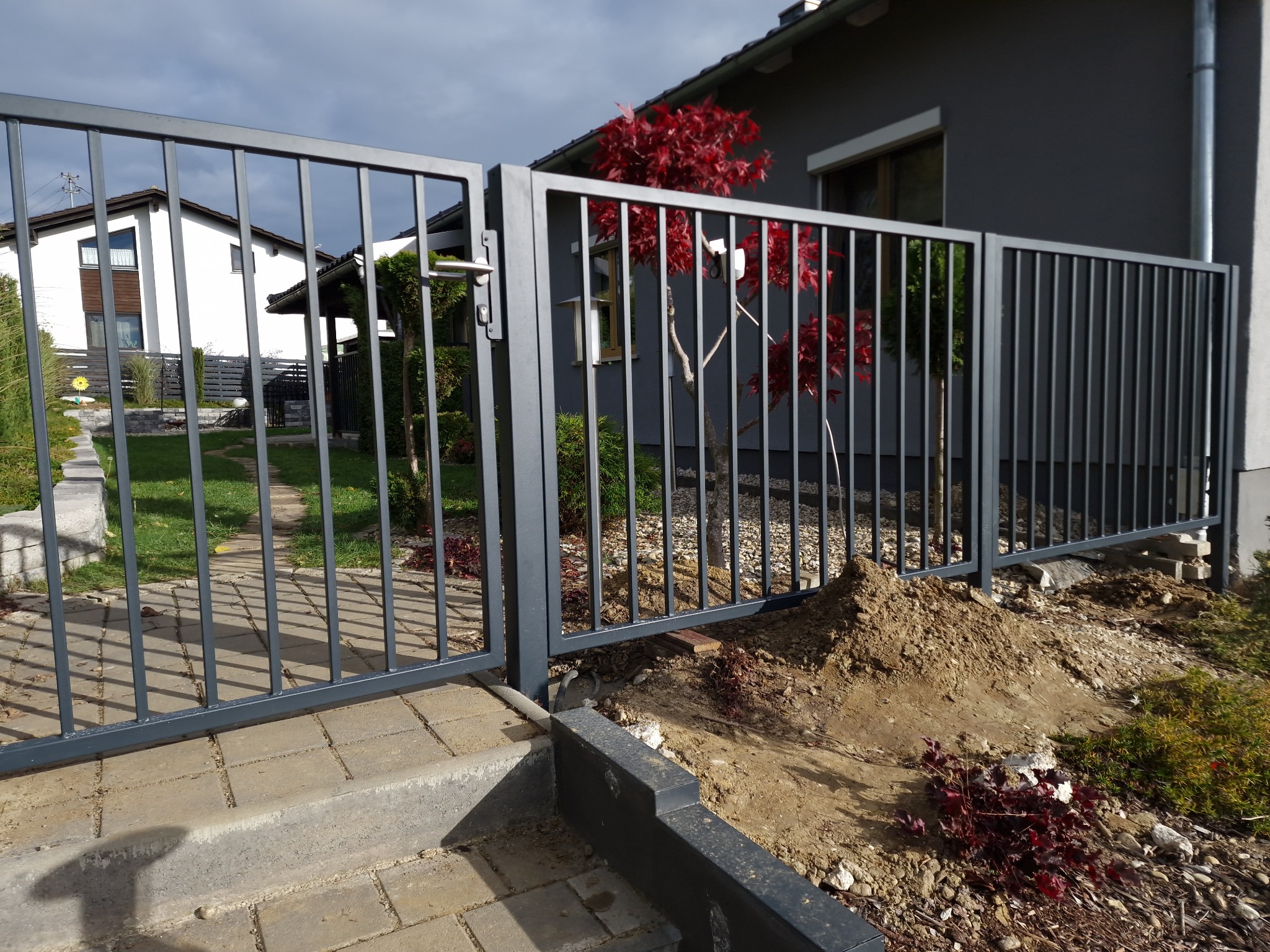 Nowoczesna, grafitowa brama wjazdowa z pionowymi szczebelkami, częściowo otwarta, prowadząca do domu z ogrodem. Widoczne świeżo nasadzone rośliny.