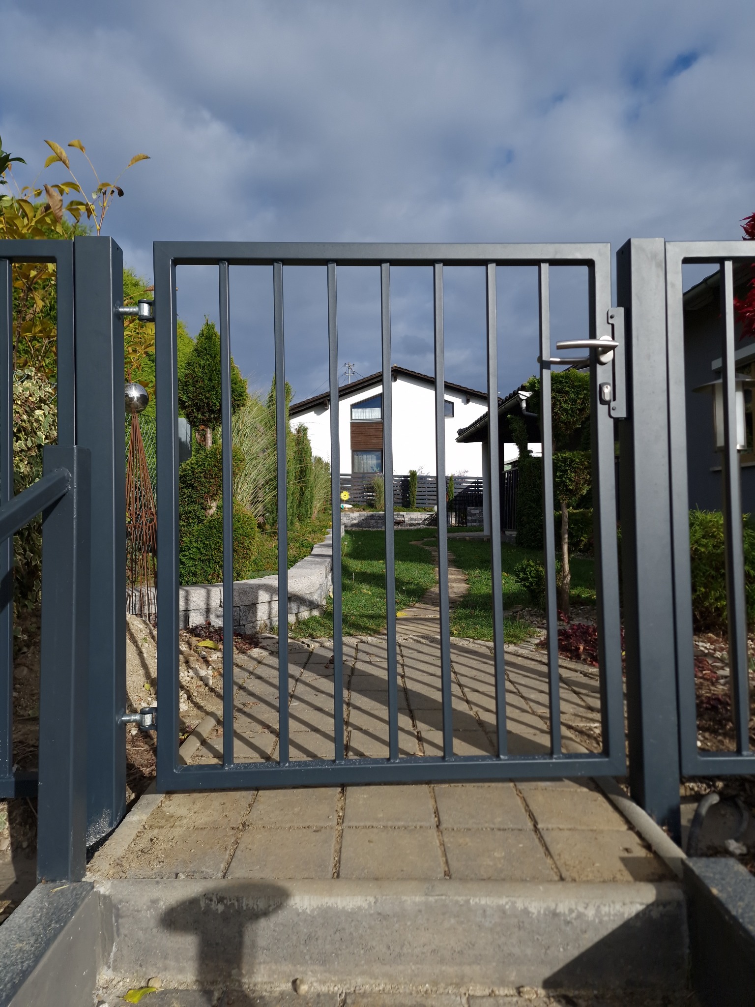 Nowoczesna, grafitowa brama wjazdowa z pionowymi szczebelkami, widziana z bliska, z domem i zielenią w tle. Minimalistyczny design, równomierne oświetlenie.