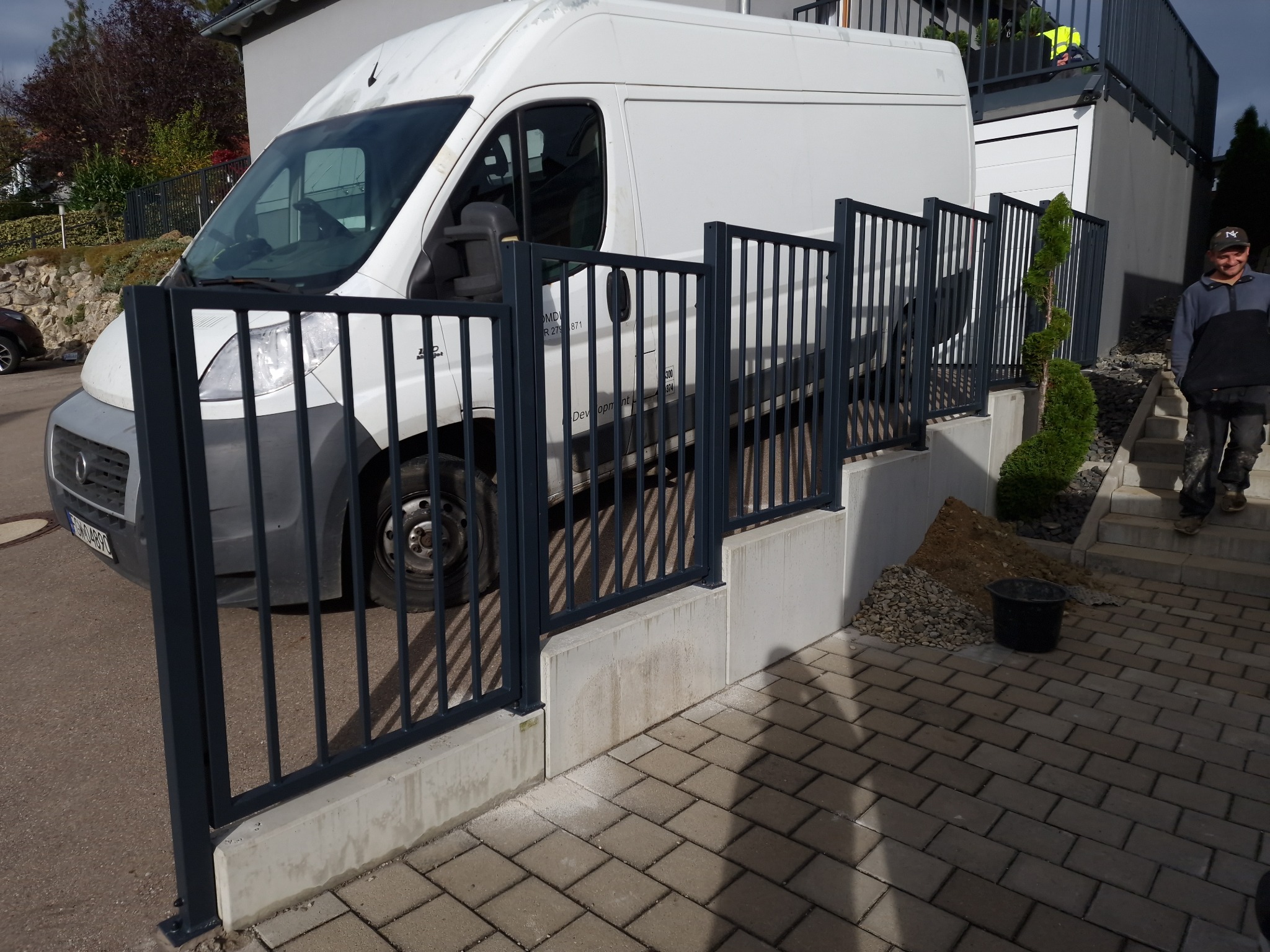 Ciemnoszare, pionowe ogrodzenie z metalowych prętów, zamontowane na betonowej podmurówce, z białym busem w tle i mężczyzną wchodzącym po schodach.