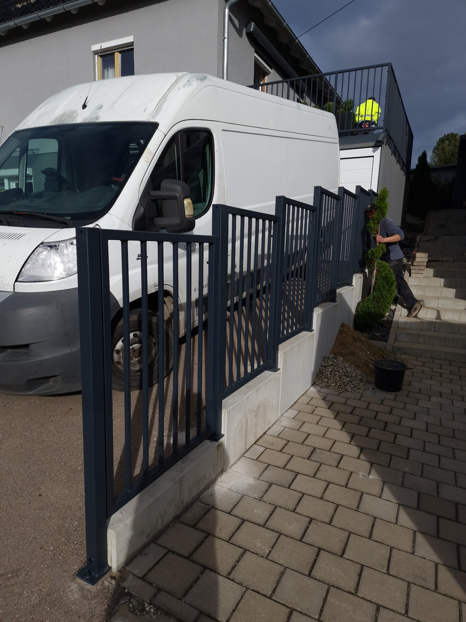 Nowoczesne, grafitowe ogrodzenie posesji z pionowych sztachet, zamontowane na betonowym murku. W tle biały bus i schody z pracownikiem w trakcie sadzenia roślin.