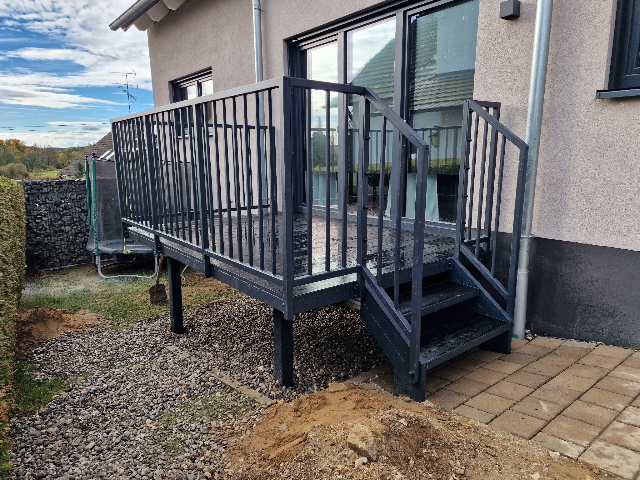 Nowoczesny, czarny taras z metalową balustradą i schodami, przylegający do domu. Widoczne kamienie i kostka brukowa wokół tarasu. Ujęcie z dołu, podkreślające konstrukcję.