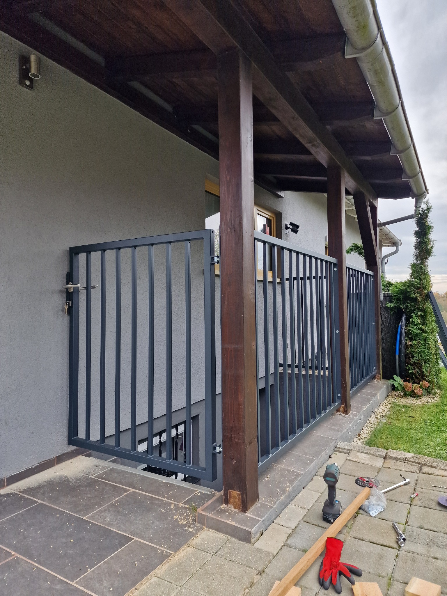 Szara, metalowa furtka z pionowymi szczebelkami, zamontowana przy wejściu na zewnątrz budynku. Widoczne narzędzia i rękawice robocze sugerują etap montażu.