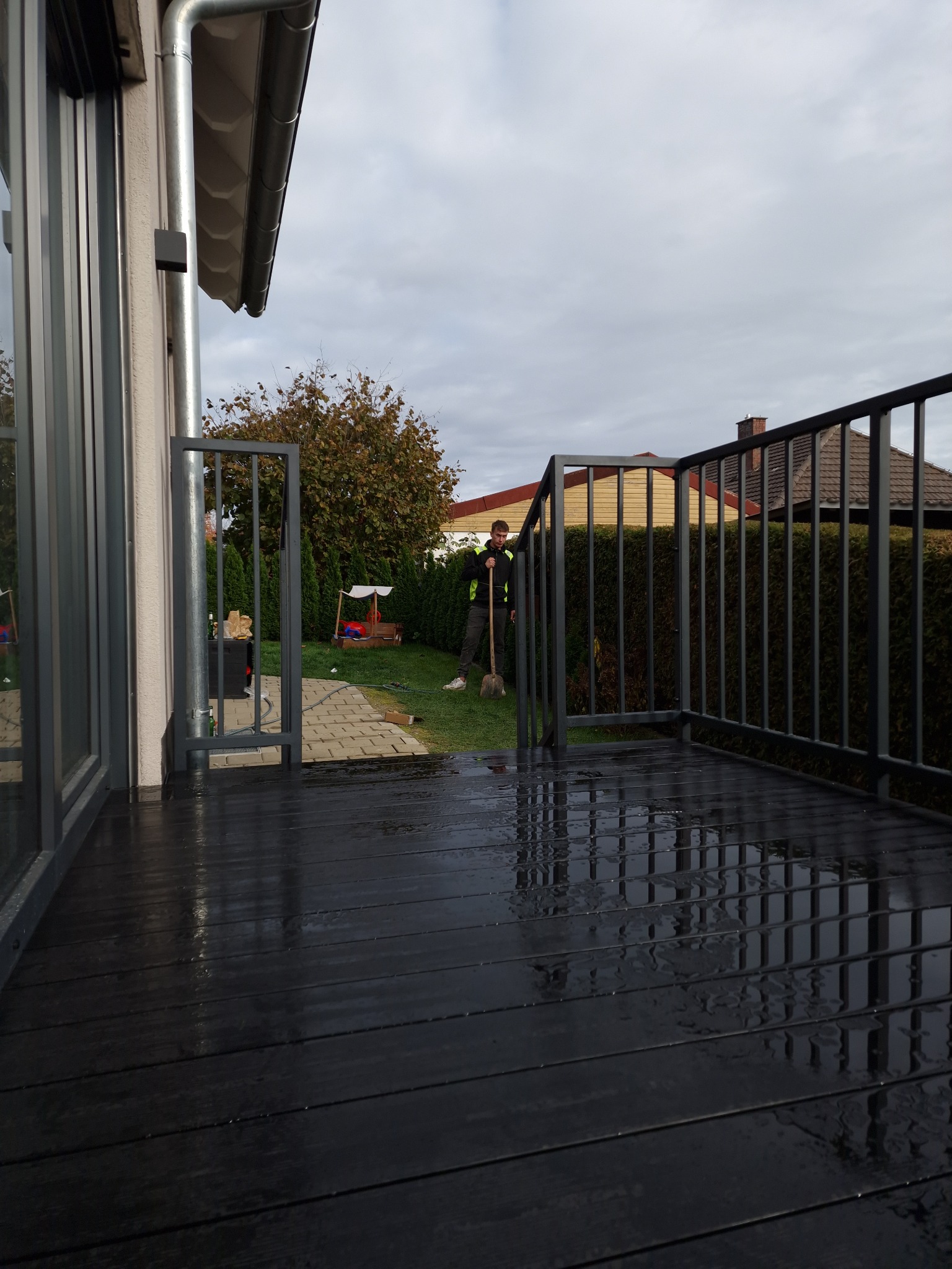 Ciemna, metalowa balustrada balkonowa z furtką, widok z góry na mokrą podłogę balkonu, w tle osoba z łopatą w ogrodzie.
