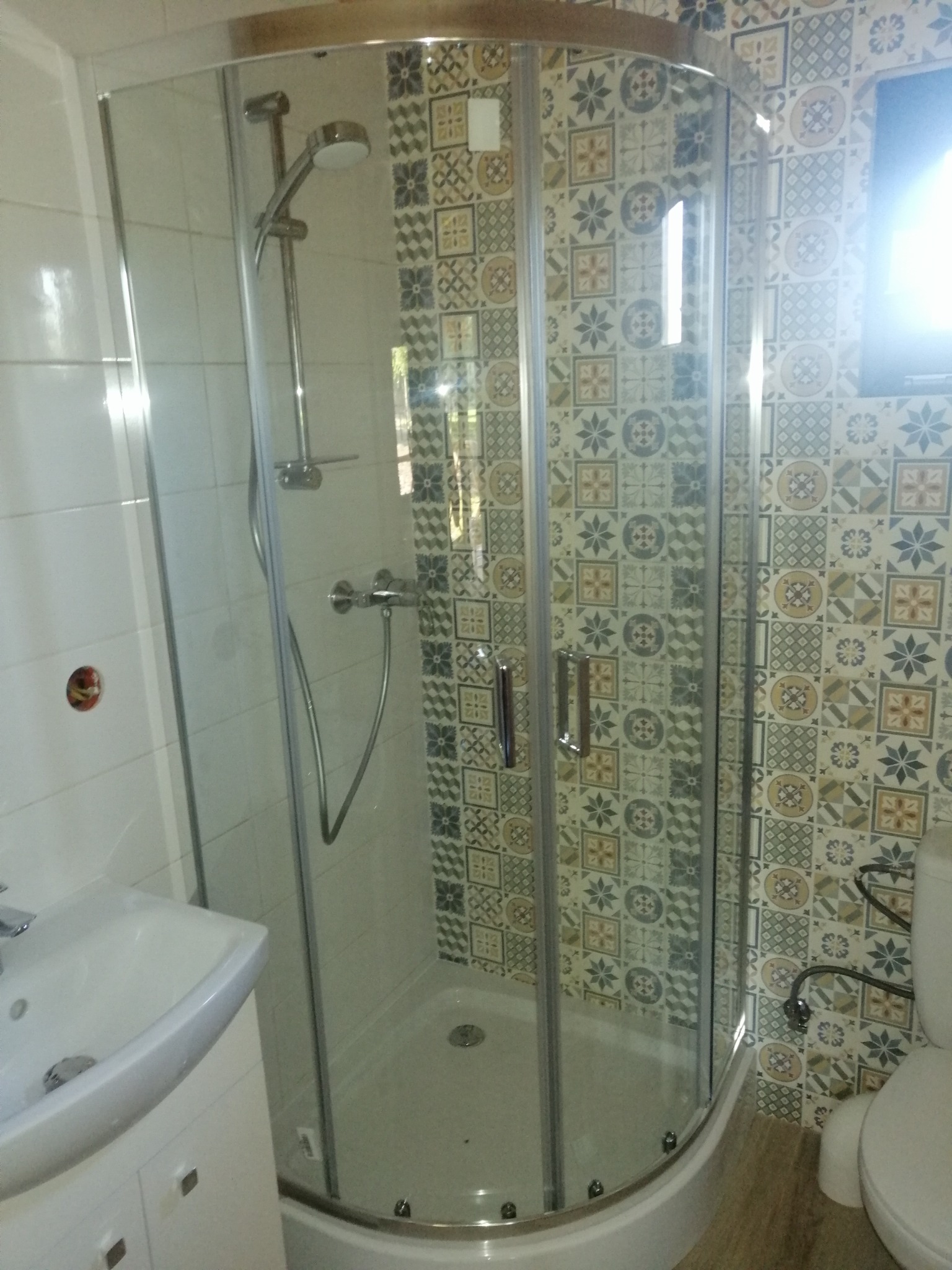 Kabinowa narożna z chromowanymi profilami i deszczownicą na tle ściany wyłożonej patchworkowymi płytkami ceramicznymi w odcieniach beżu, szarości i błękitu, widoczna umywalka i toaleta.