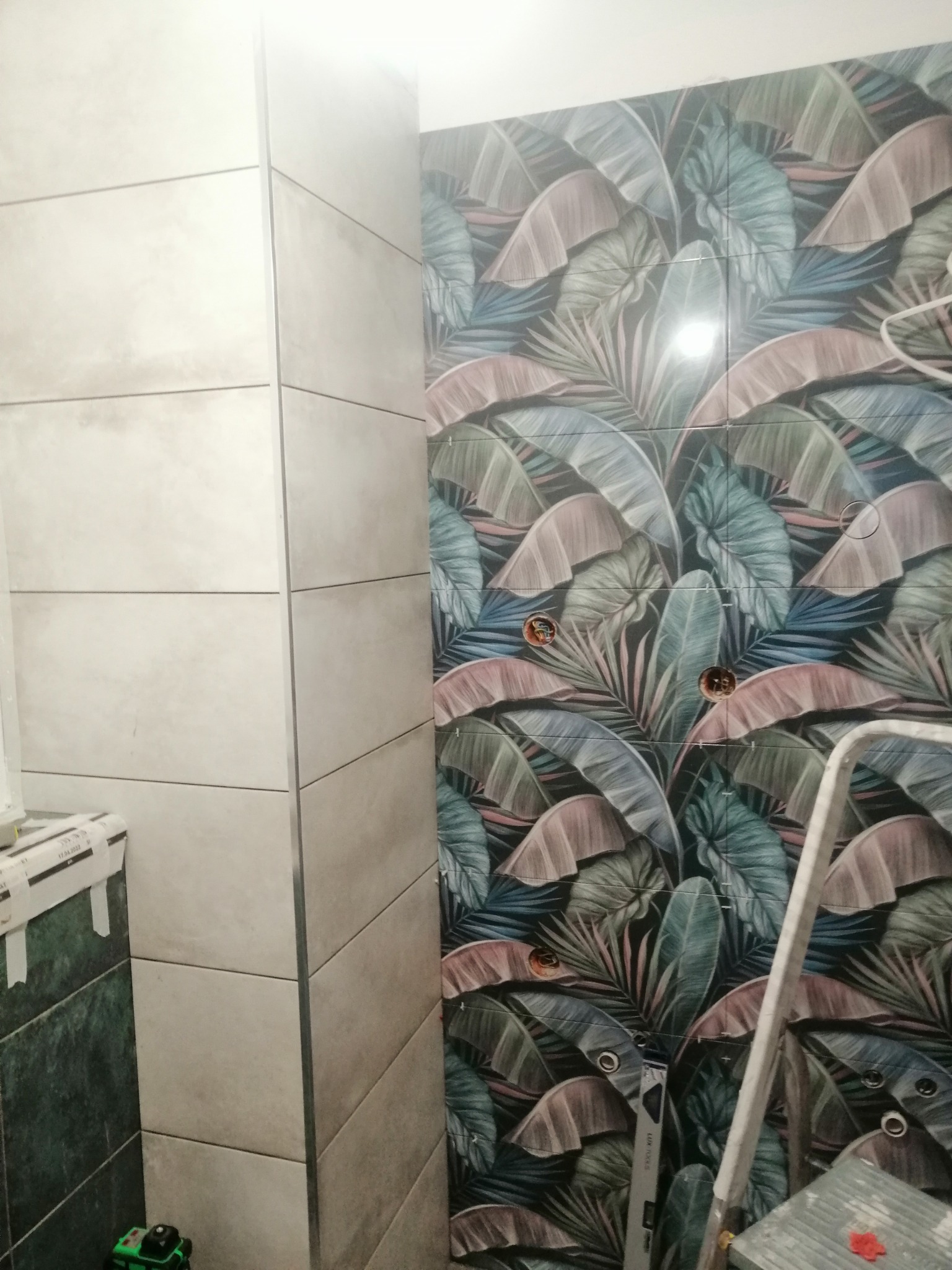 Ściana łazienki wyłożona płytkami: z lewej strony szare, poziome płytki, z prawej płytki z motywem liści, widoczne otwory pod instalacje, drabina oparta o ścianę.