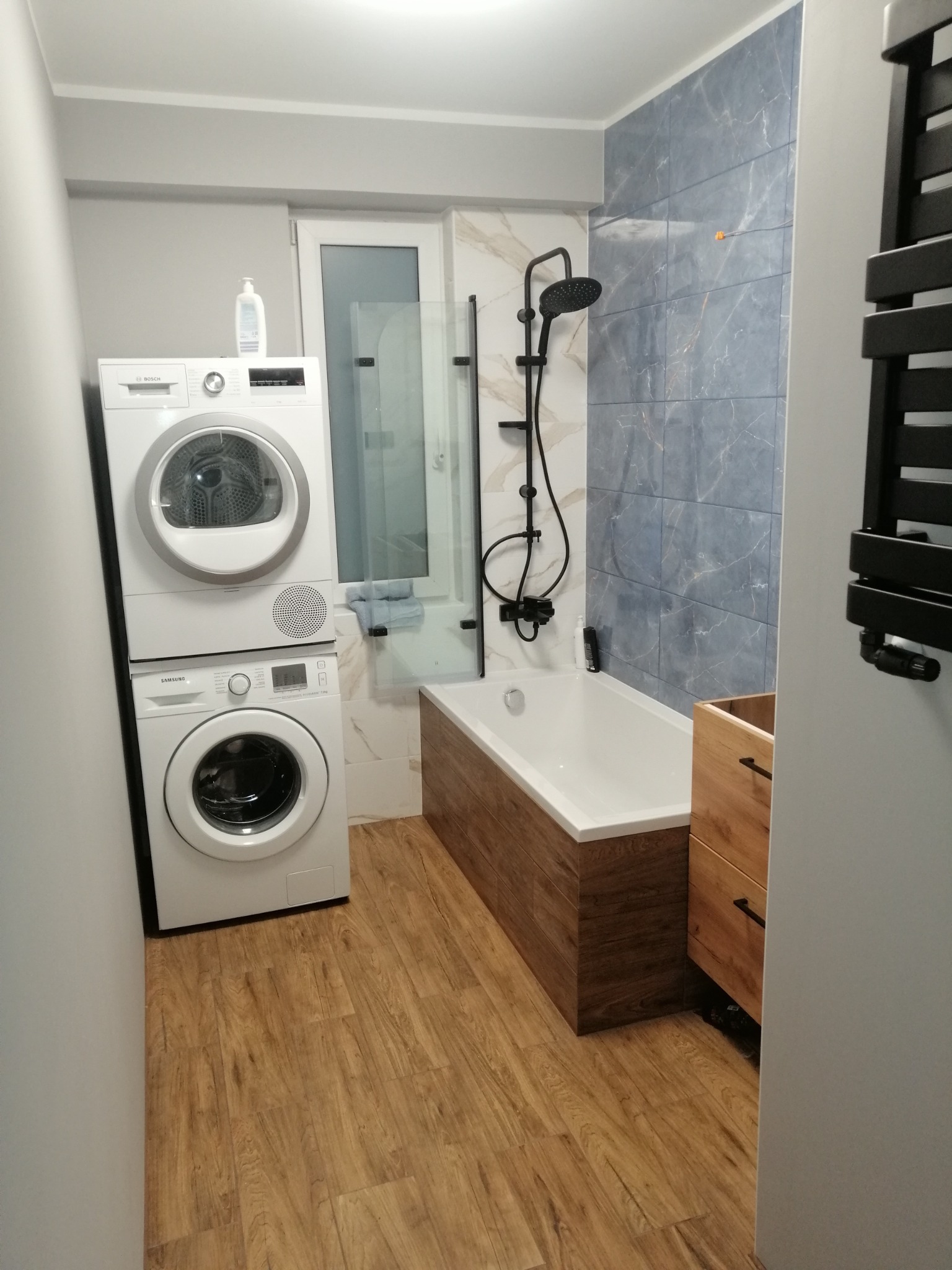 Nowoczesna łazienka z wanną obudowaną panelami imitującymi drewno, ciemną armaturą, niebiesko-szarymi płytkami na ścianie, pralką i suszarką ustawionymi jedna na drugiej oraz drewnopodobną podłogą.