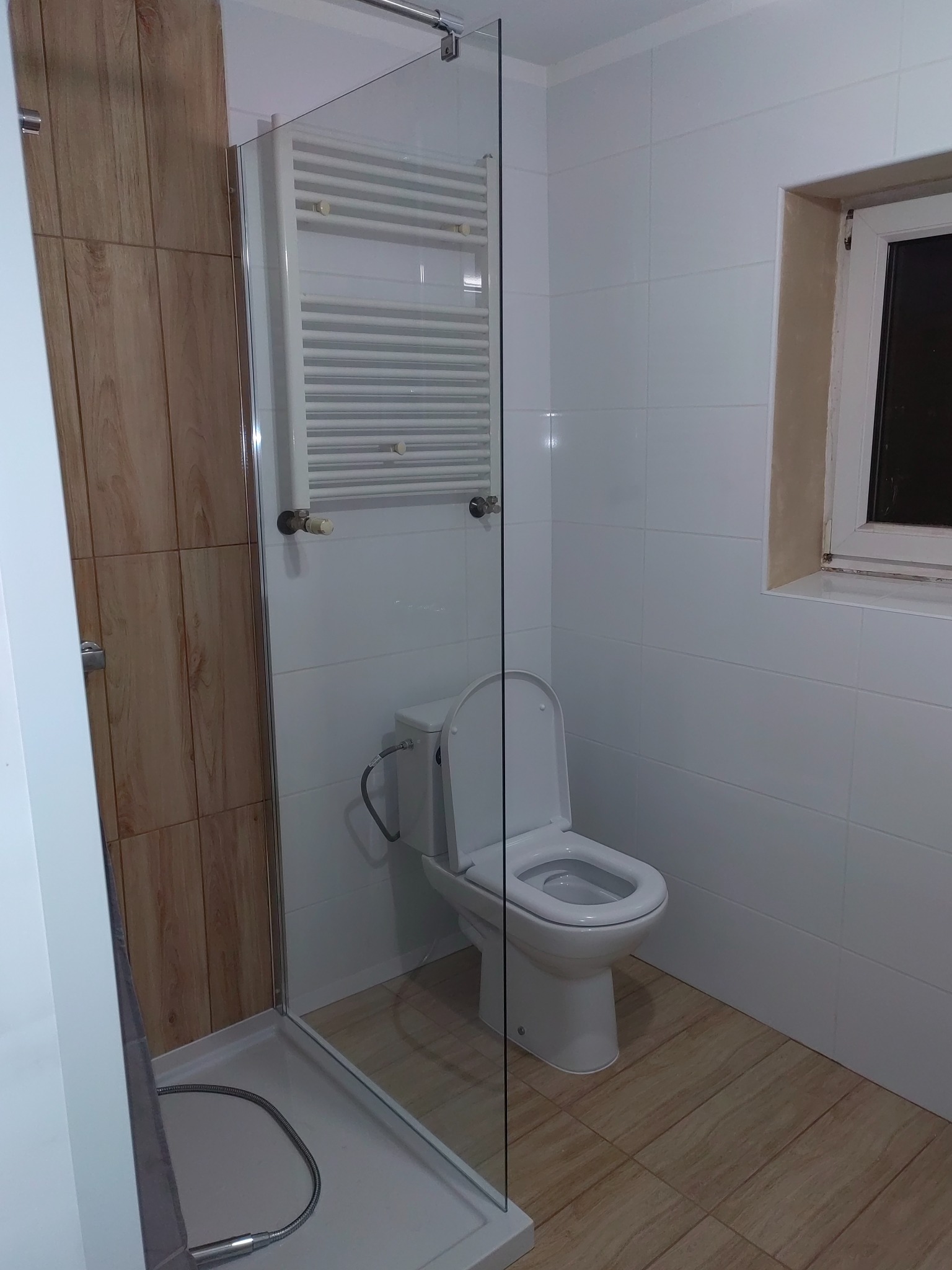 Wykończona łazienka z kabiną prysznicową ze szklaną ścianką, wiszącym grzejnikiem drabinkowym i toaletą w kolorze białym, na podłodze płytki imitujące drewno.