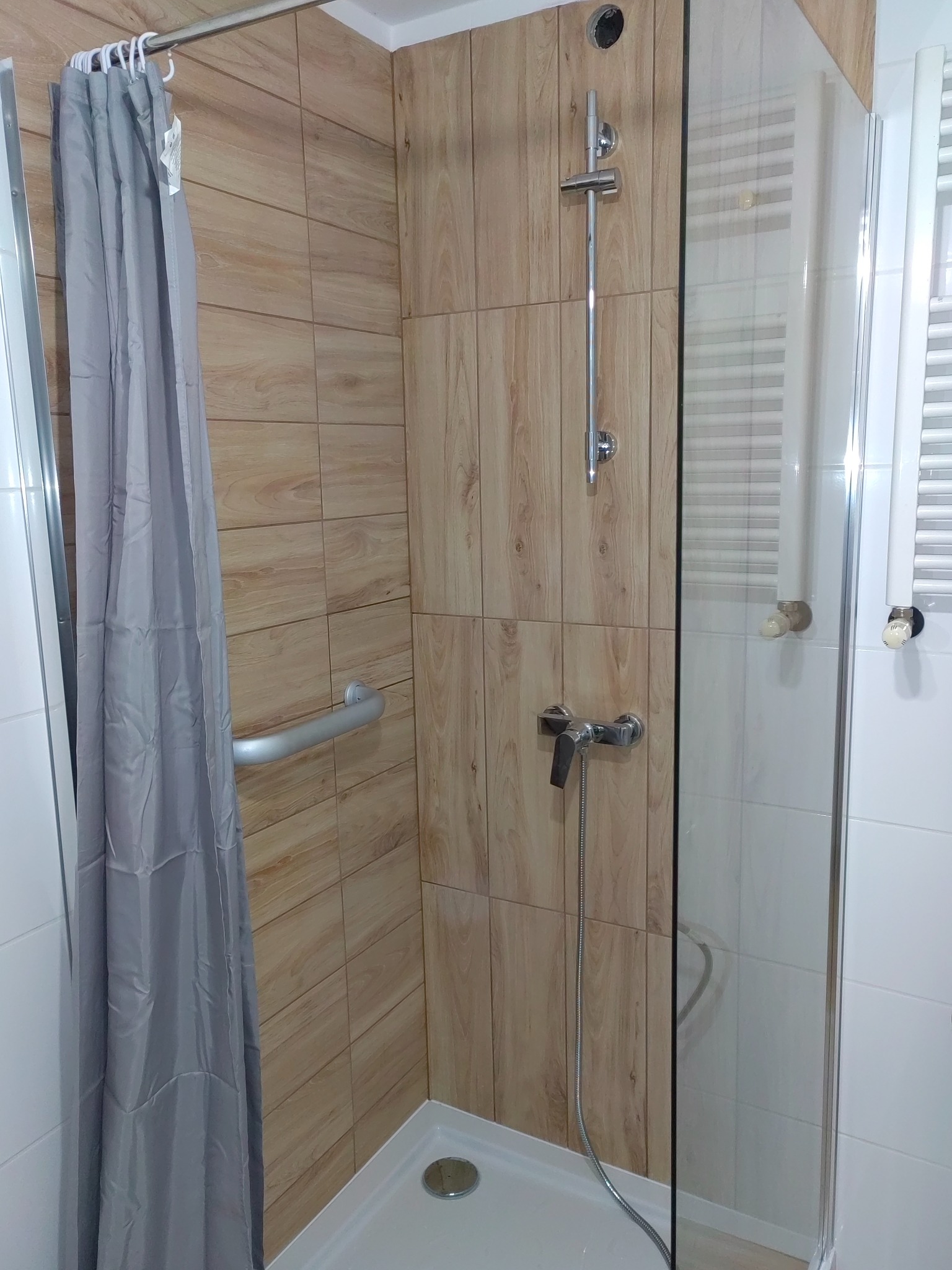 Kabina prysznicowa z szarym zasłonką, drewnopodobne płytki na ścianie, uchwyt dla niepełnosprawnych i biały grzejnik drabinkowy.