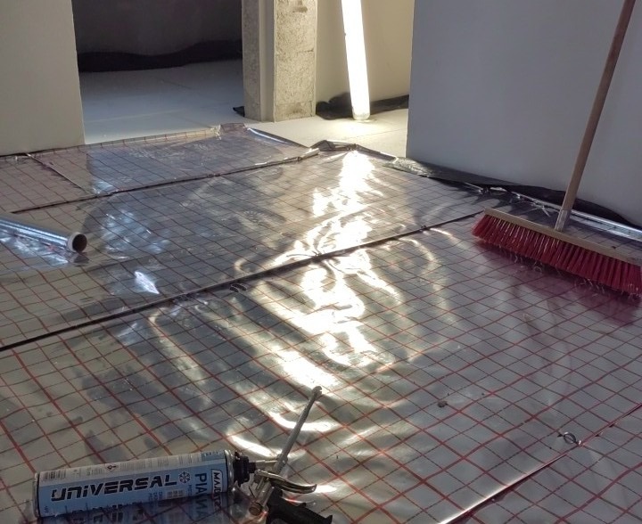 Przygotowanie podłoża pod system grzewczy: folia aluminiowa z siatką, tuba z pianką montażową i szczotka na podłodze w surowym wnętrzu.