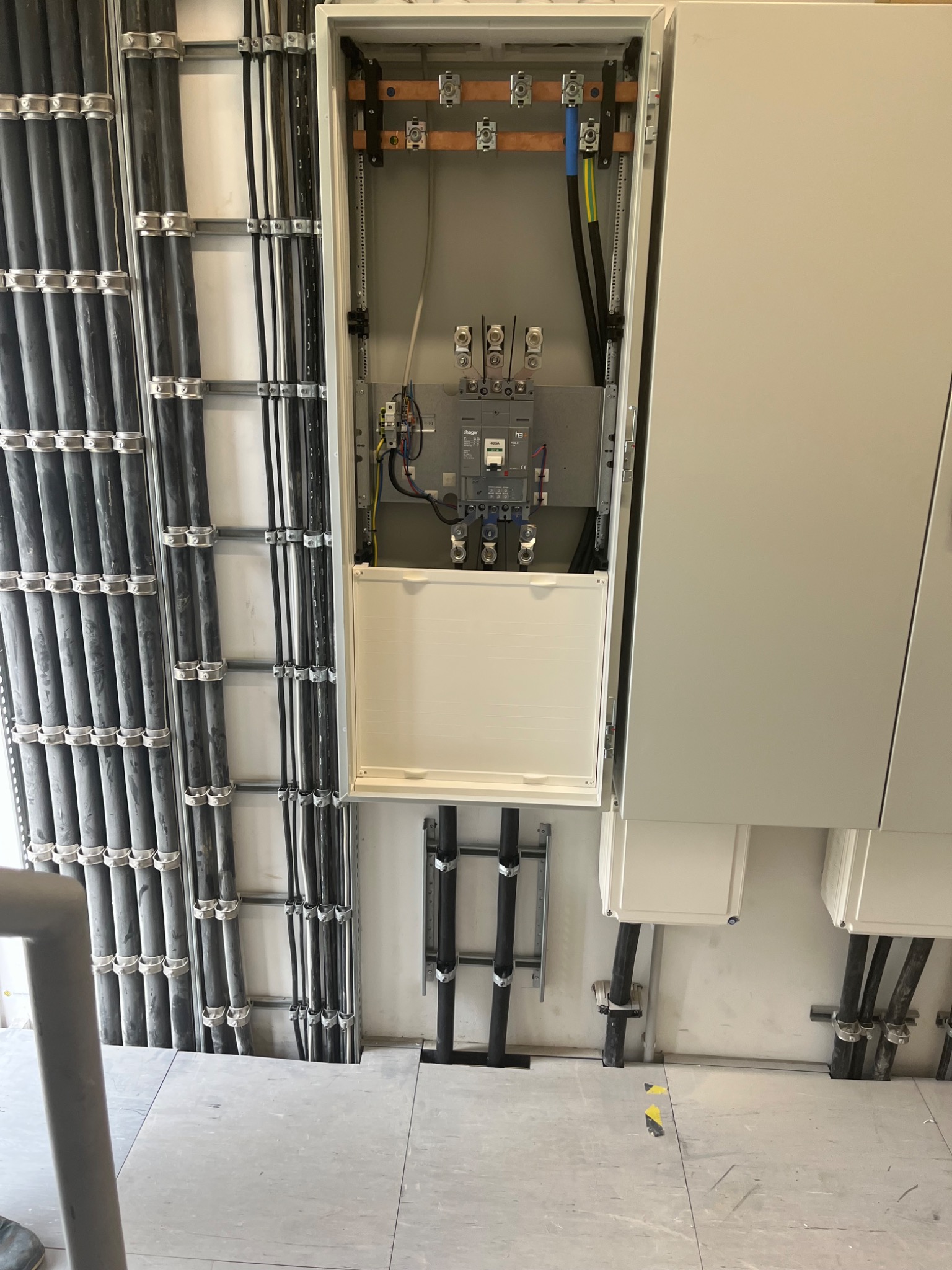 Otwarta skrzynka rozdzielcza z widocznymi bezpiecznikami i przewodami, liczne kable w peszlach przymocowane do ściany za pomocą metalowych uchwytów.