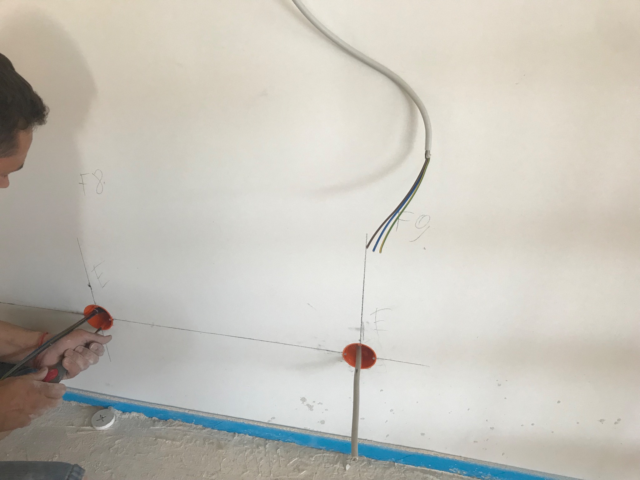 Elektryk przy ścianie z wyprowadzonym okablowaniem, zaznaczonymi punktami i puszkami instalacyjnymi, używa szczypiec do montażu.