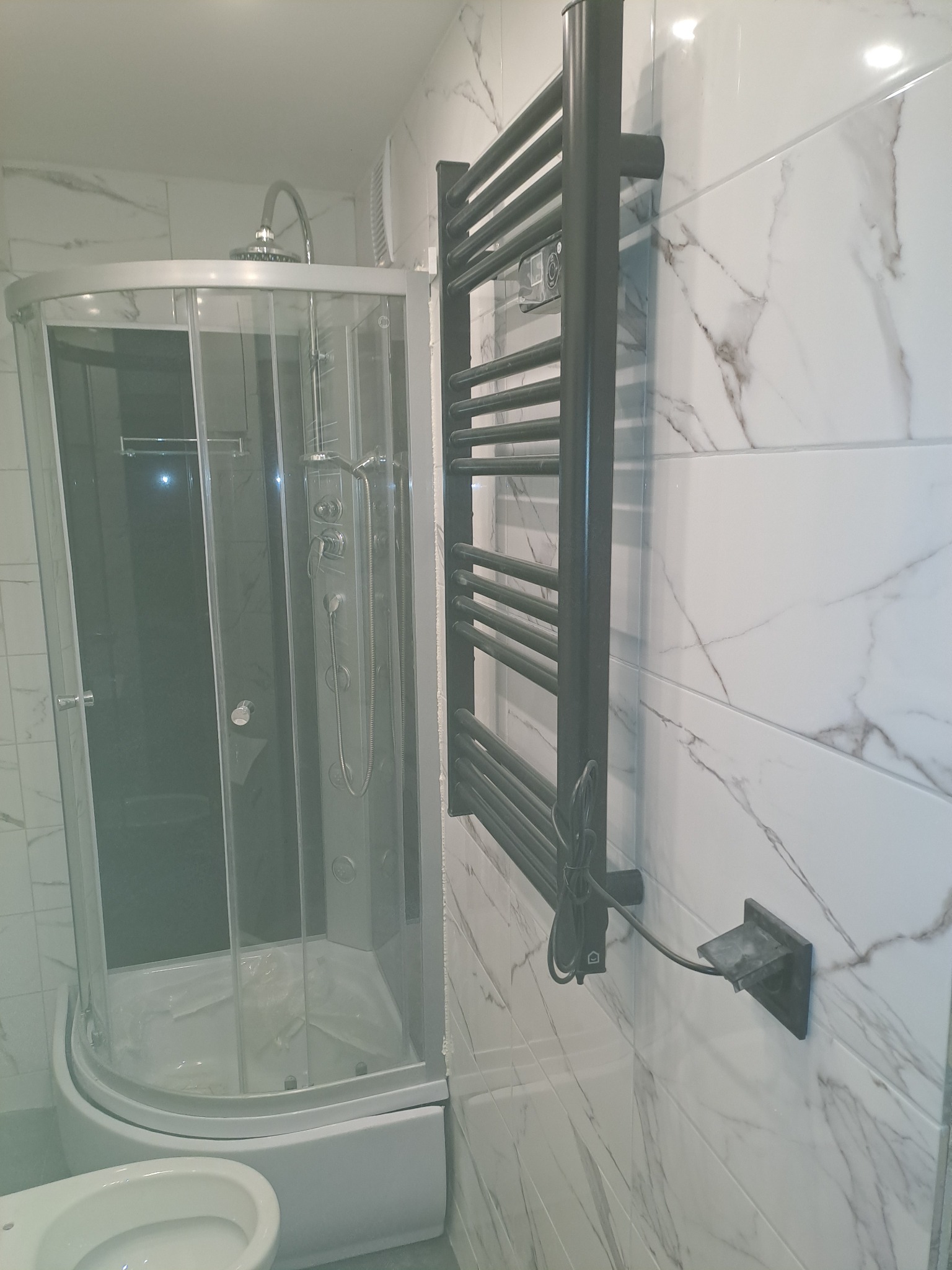 Nowoczesna kabina prysznicowa z hydromasażem, biała ceramika sanitarna i grafitowy grzejnik drabinkowy na ścianie wyłożonej marmurkowymi płytkami.